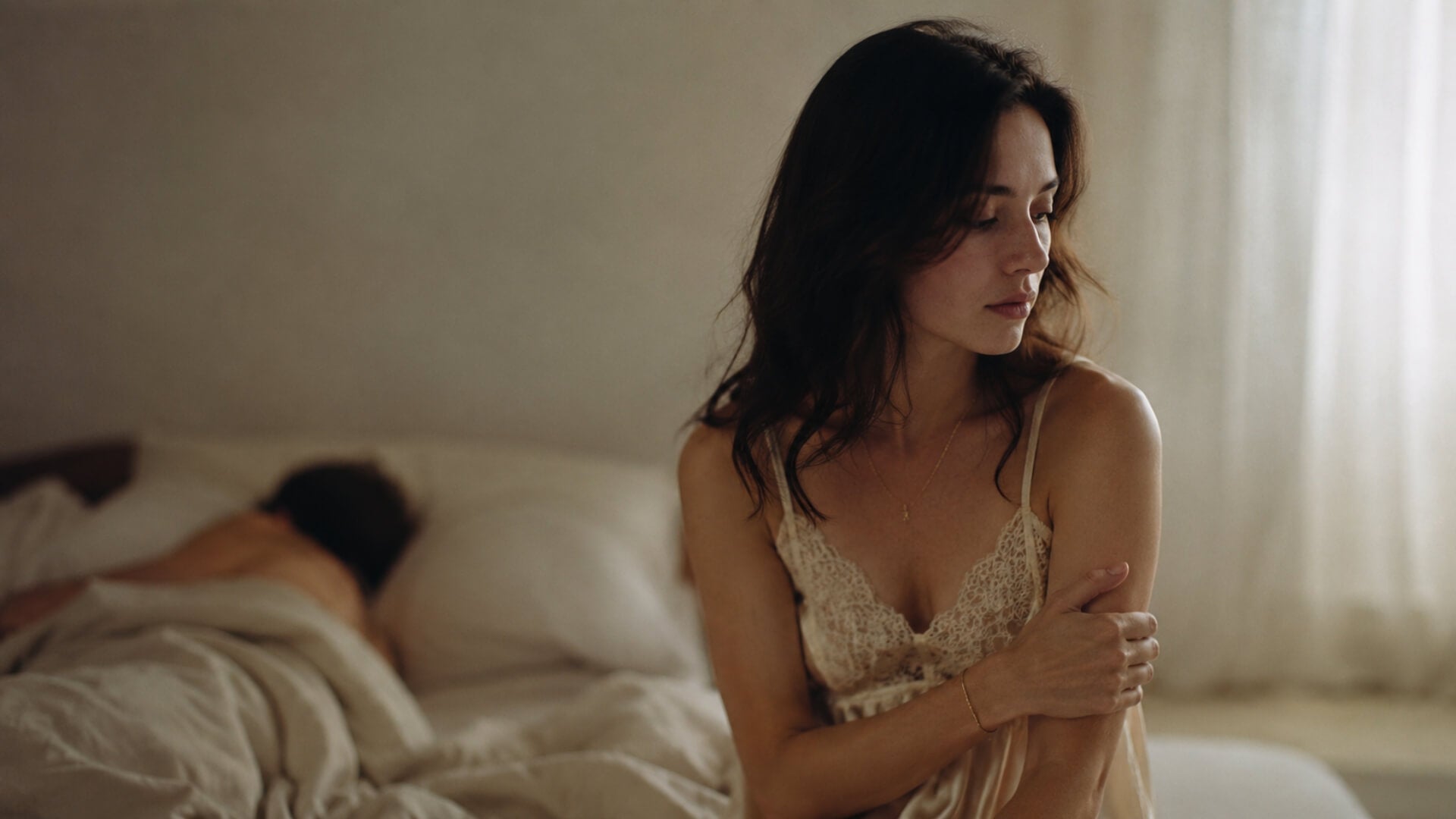 Femme assise au bord du lit, bras repliés contre elle et regard baissé, son partenaire endormi derrière elle — baisse de libido chez la femme