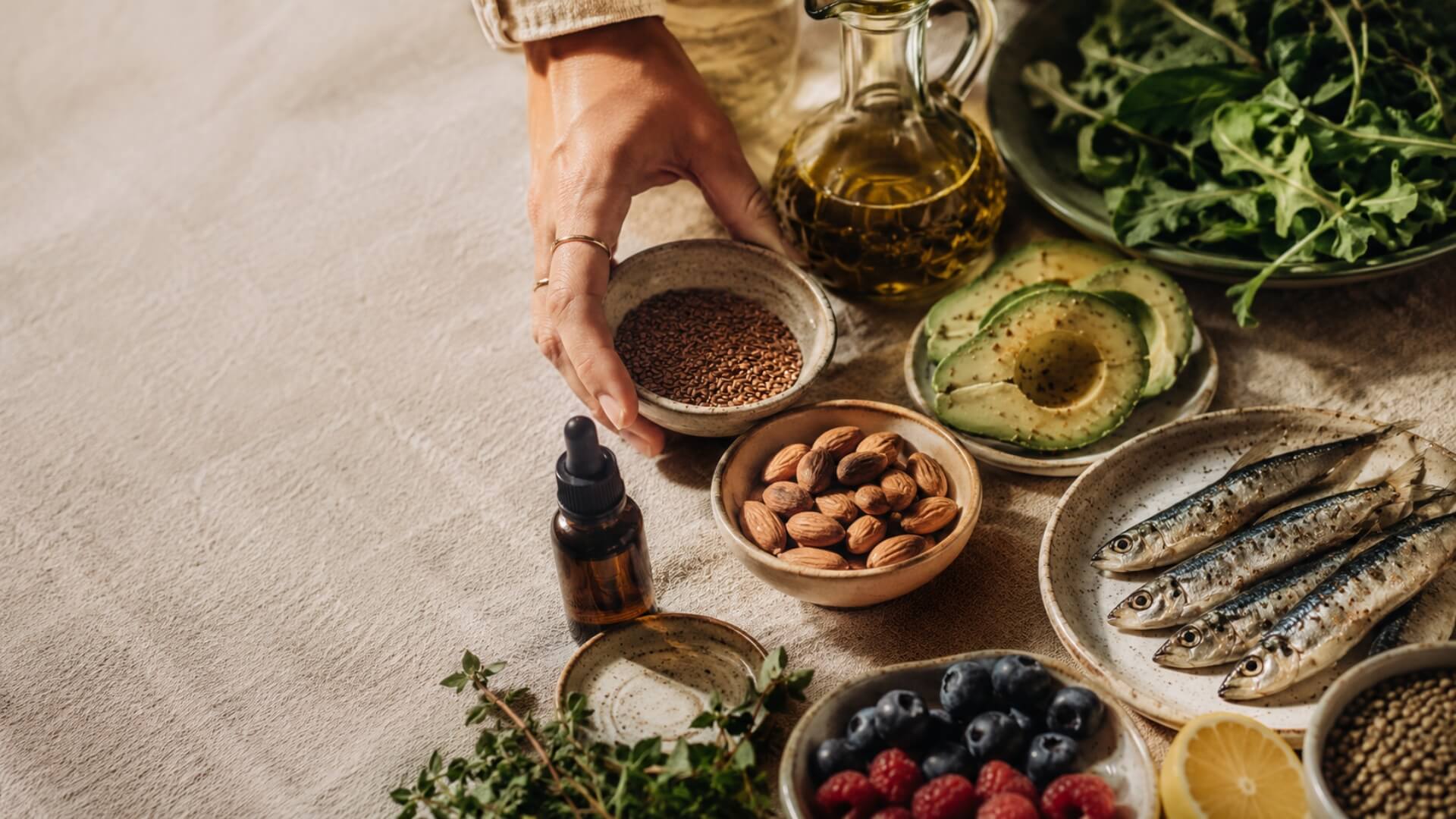Main saisissant un bol de graines parmi des amandes, un avocat, des baies rouges, de l'huile d'olive et des herbes fraîches — alimentation et compléments à privilégier en cas de SOPK
